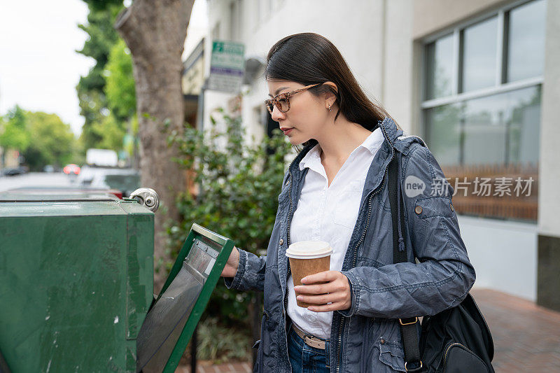 asian chinese female business person carrying coffee and opening green newspaper rack to check if thereâs still free tabloid to get on her way to work in the morning in palo alto city California usa